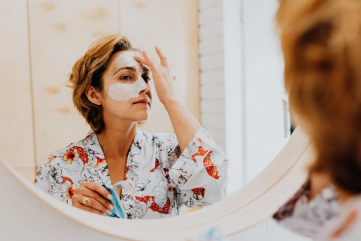 Mujer aplicándose una mascarilla facial blanca frente a un espejo redondo
