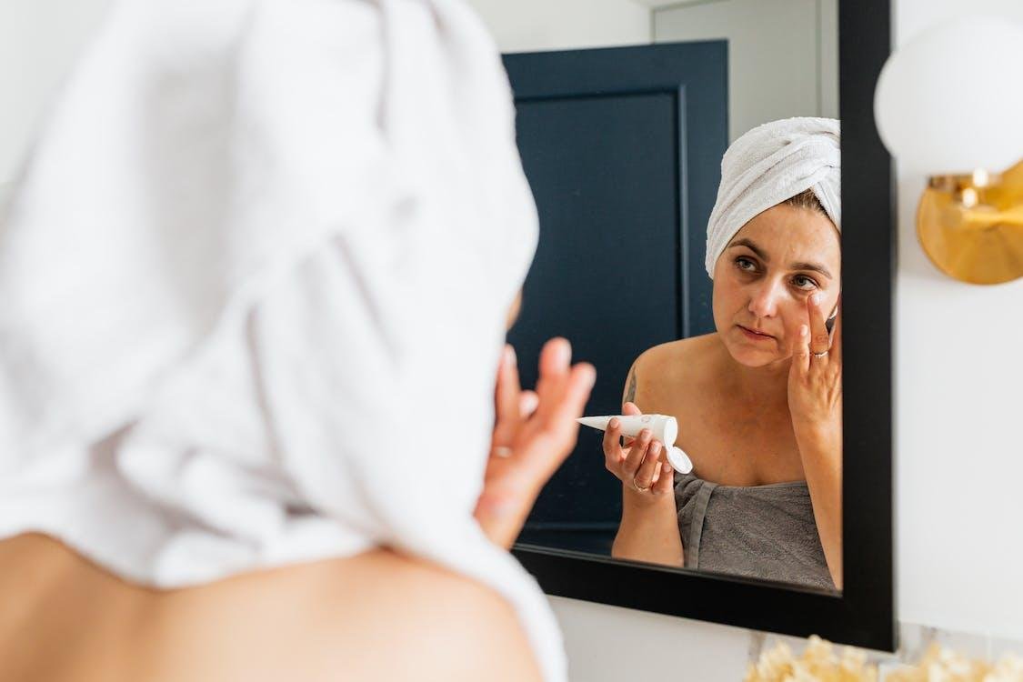 Mujer con toalla en el cabello aplicándose un producto en el contorno de ojos frente al espejo
