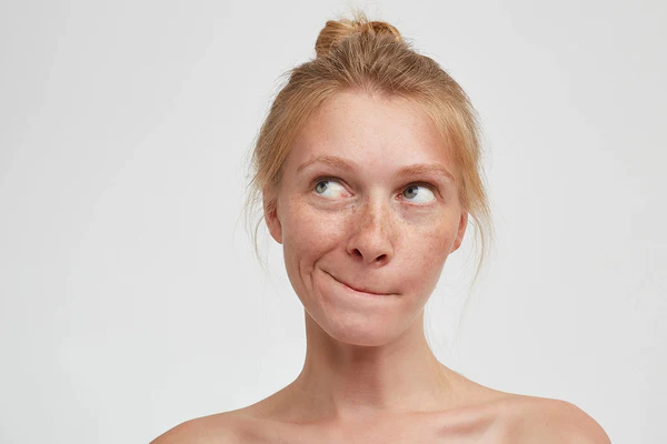 Mujer pelirroja con pecas en el rostro y expresión de duda sobre fondo claro