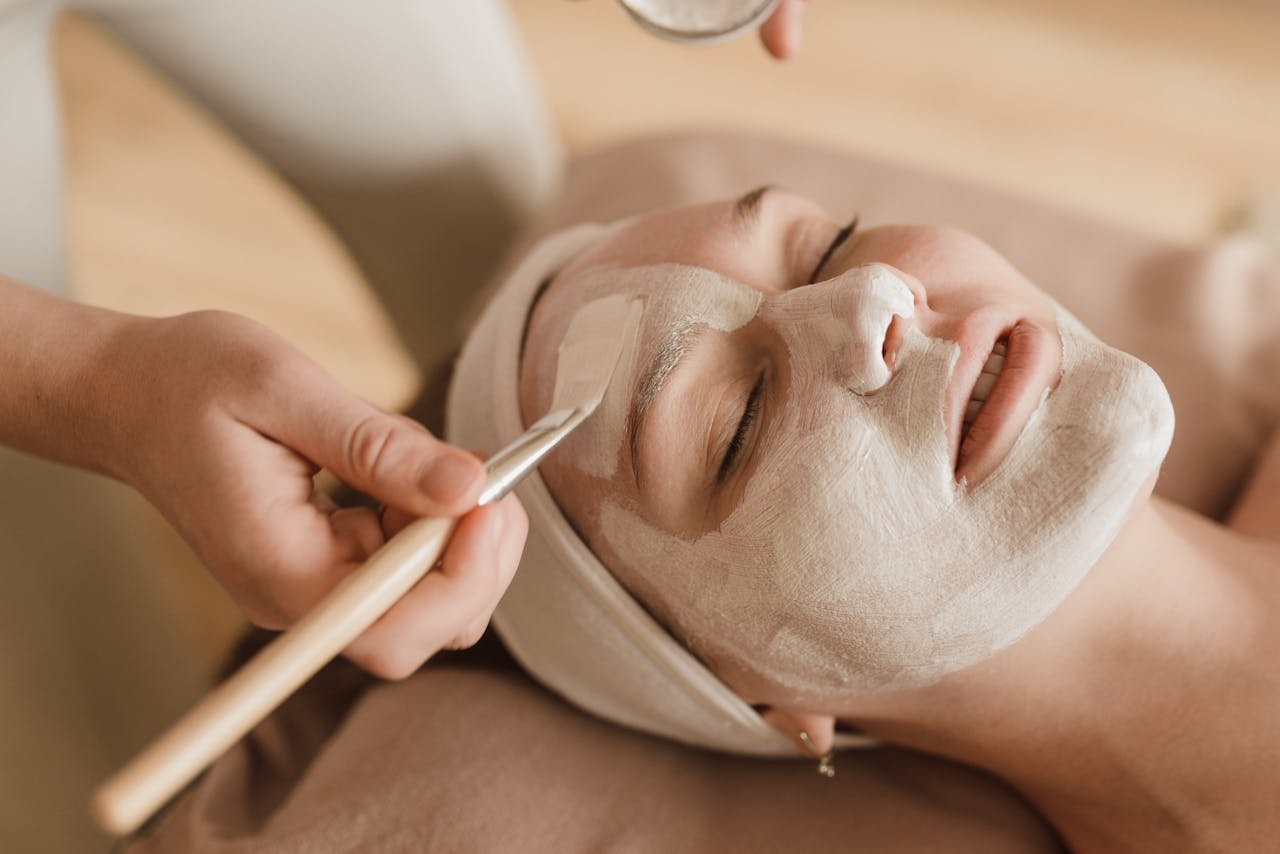 Mujer recostada con los ojos cerrados mientras le aplican una mascarilla facial de arcilla con pincel en un tratamiento profesional