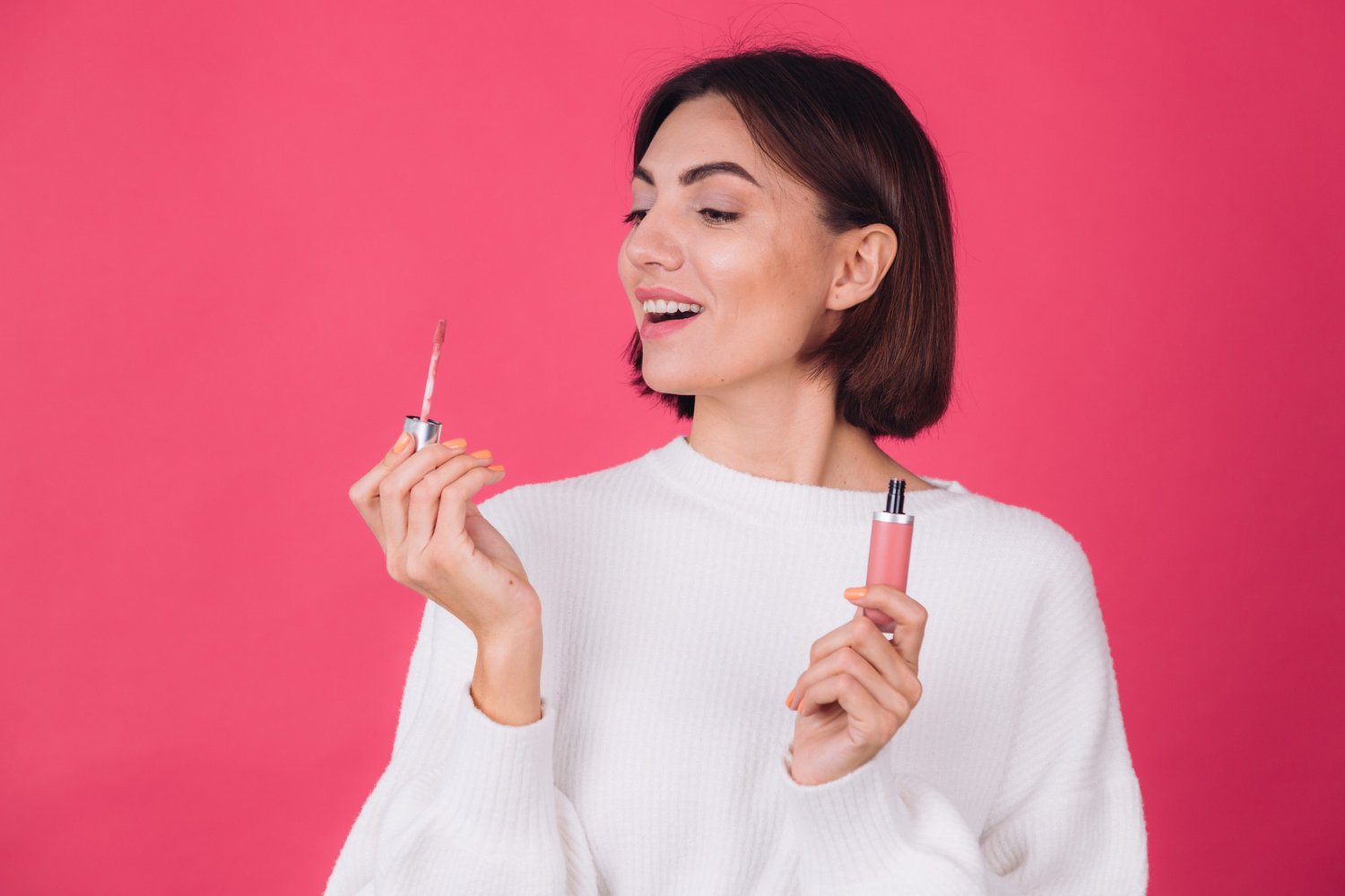 Mujer sonriente con un labial voluminizador en cada mano sobre fondo rosado