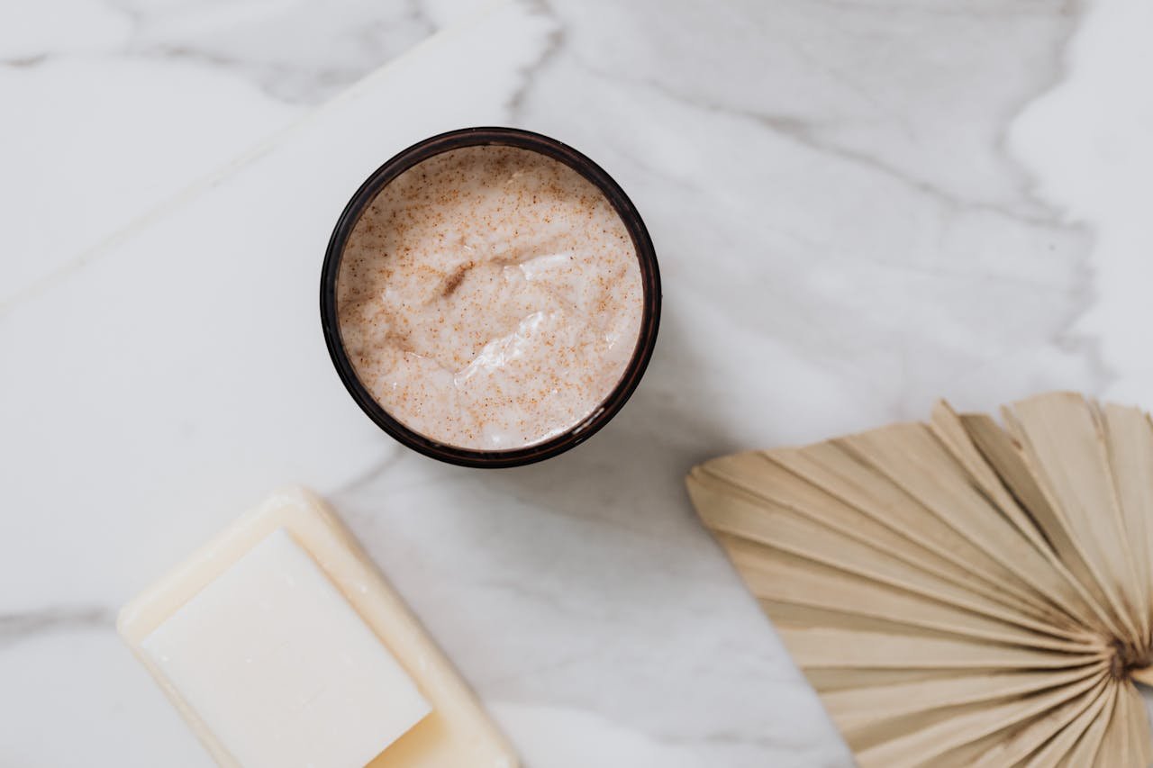 Open jar of grainy-textured face cream on white marble next to a paper fan and a bar of soap
