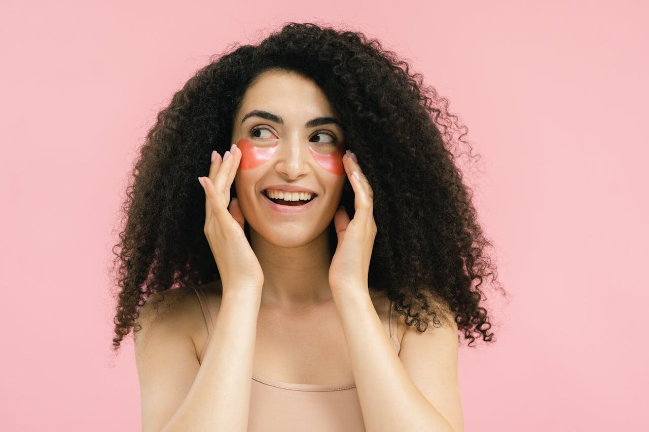 Smiling woman with curly hair wearing hydrating under-eye patches against a pink background