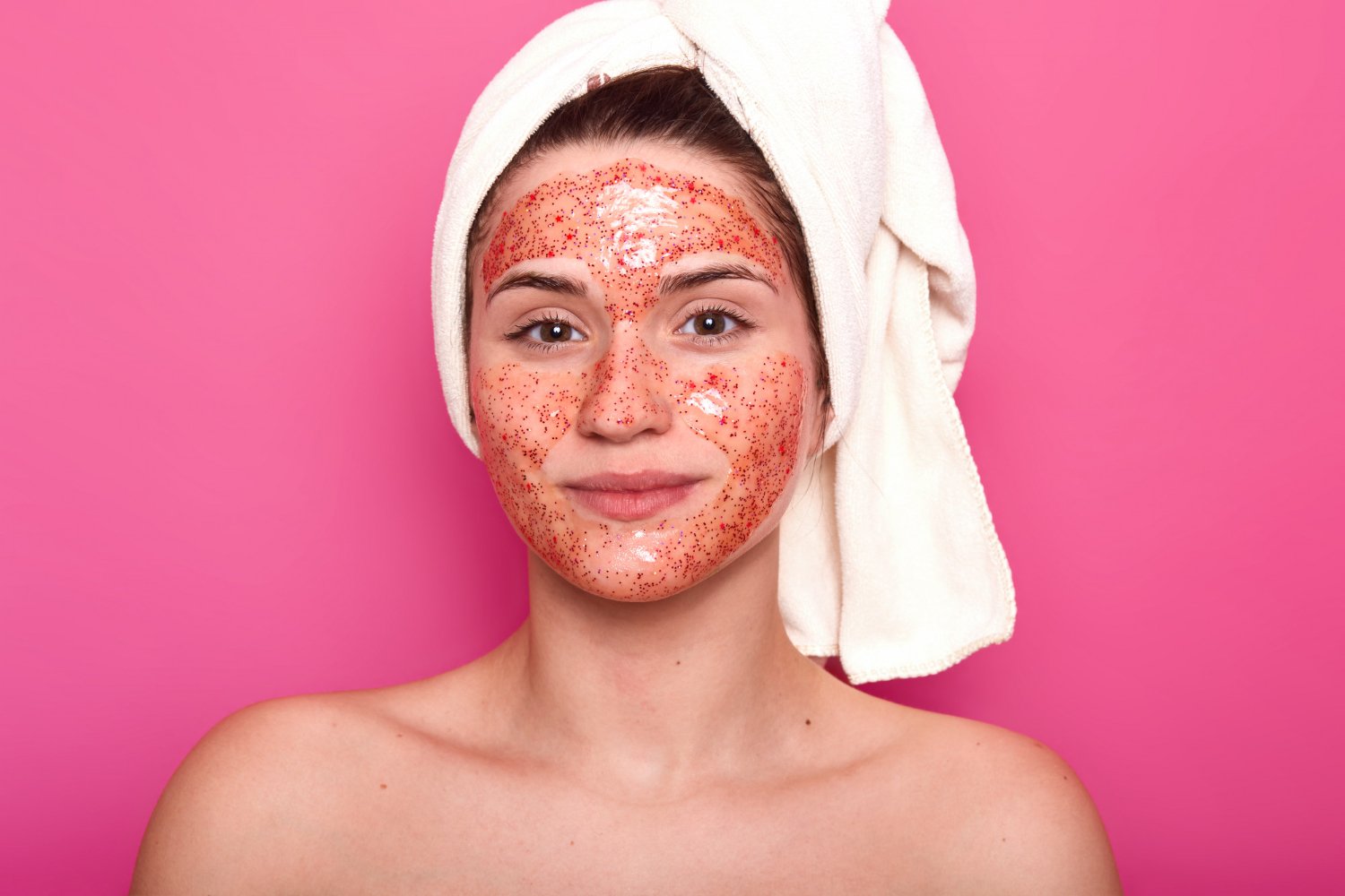 Mujer con toalla en el cabello y una mascarilla facial roja aplicada en todo el rostro sobre fondo rosado