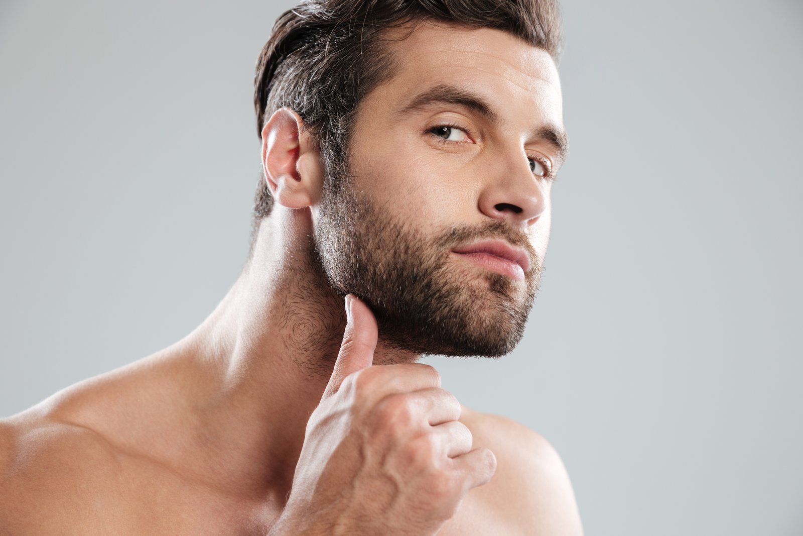 Hombre con barba poblada se toca la mandíbula mientras revisa su rostro sobre fondo gris