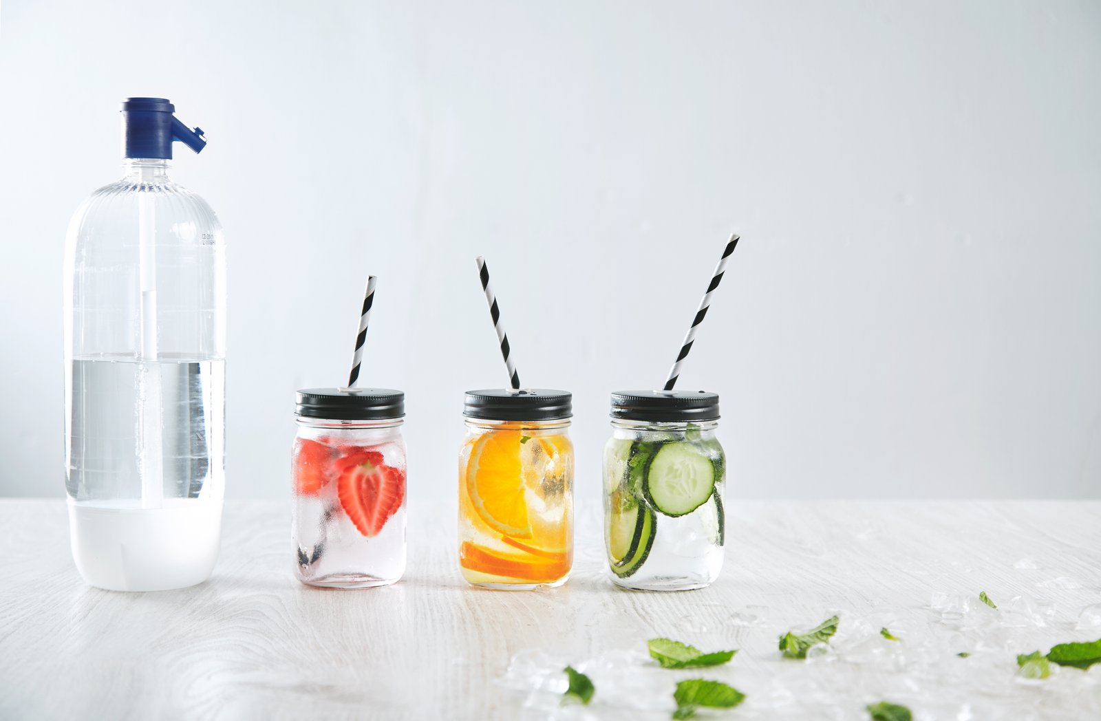 Botella de agua con gas junto a tres frascos con agua de fresa, naranja y pepino